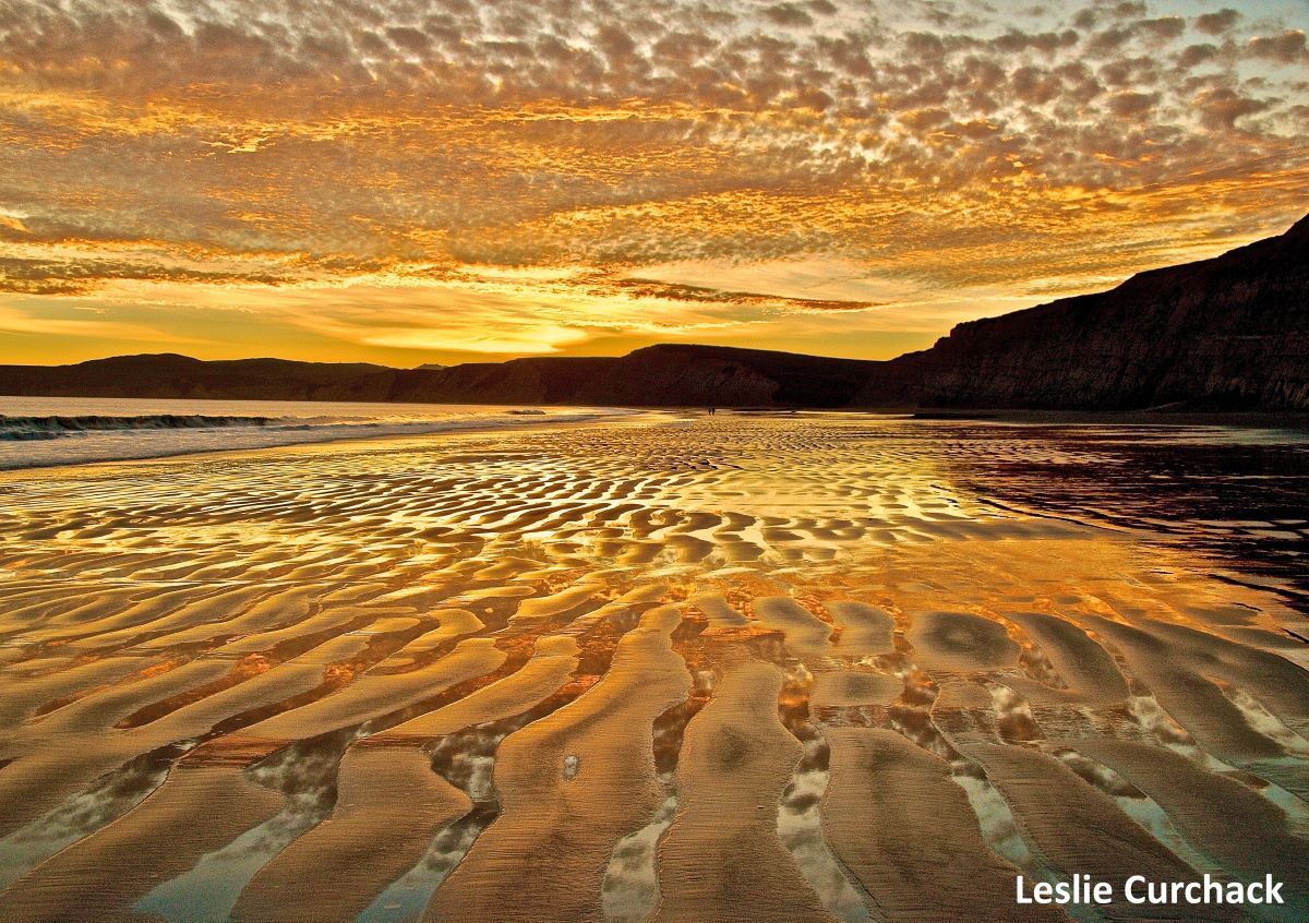 A bright orange sunset and scattered clouds are reflected in the rippled patterns on the sand.